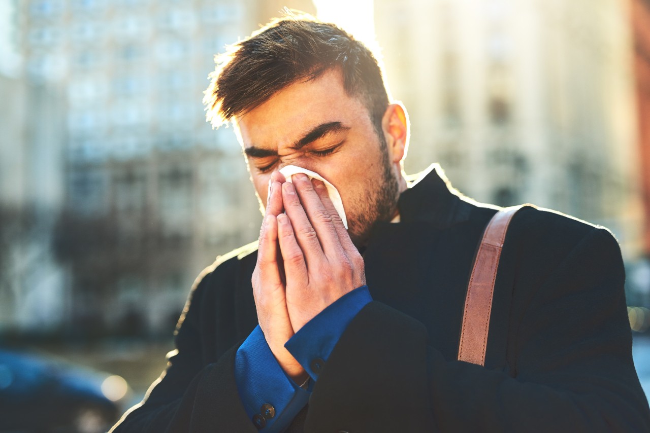 Shot of a irritated looking young man blowing his nose with a tissue while walking the busy streets of the city in the morning