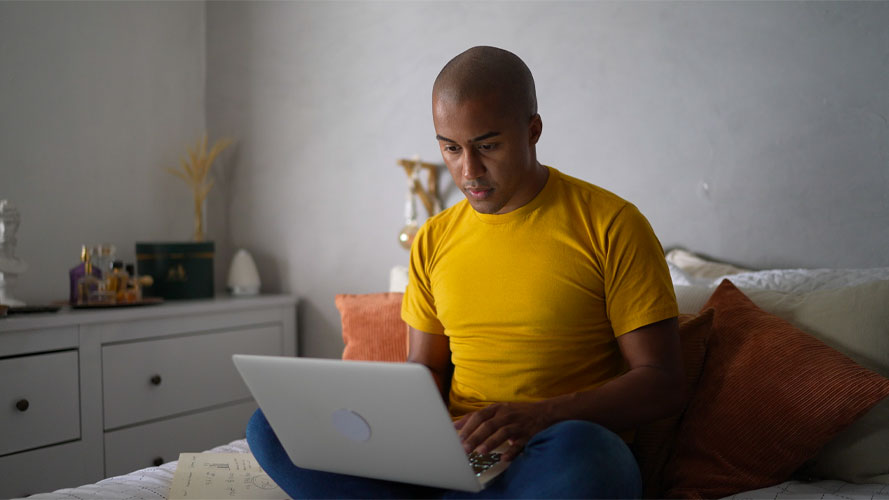 young man of color in yellow shirt uses computer sitting on bed