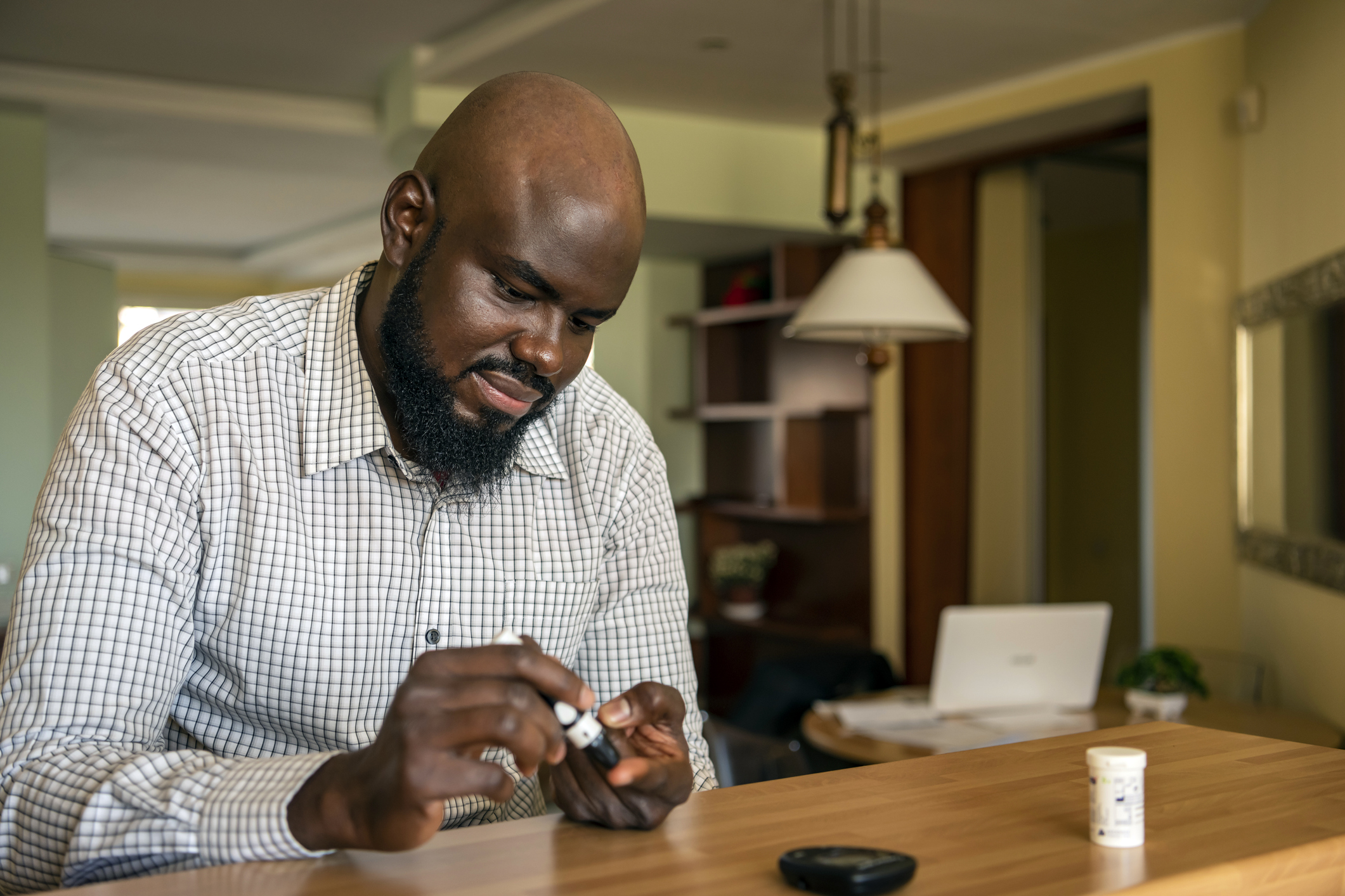 A modern African businessman is sitting at the desk at the home office and taking blood from his finger due to diabetes. The daily life of a man of African-American ethnicity person with a chronic illness who is using glucose tester.