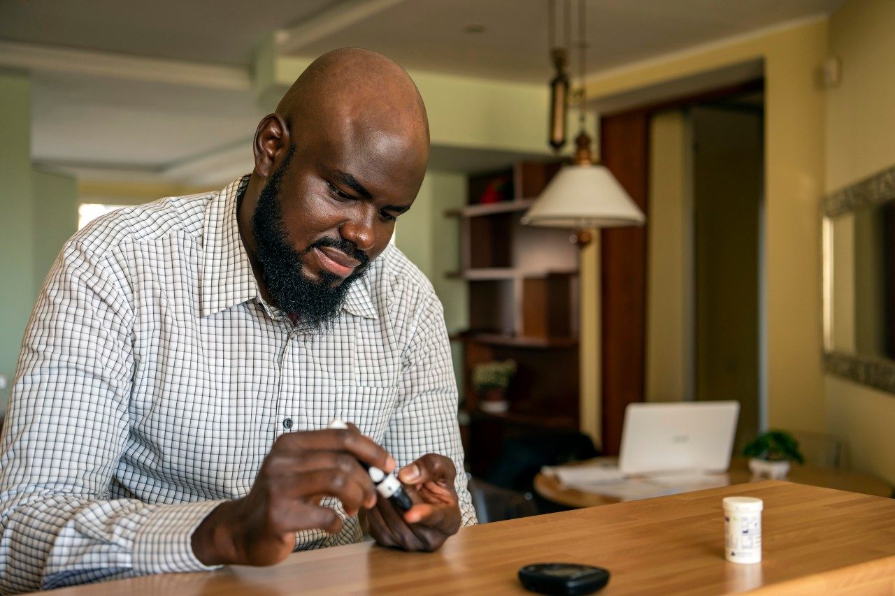 A modern African businessman is sitting at the desk at the home office and taking blood from his finger due to diabetes. The daily life of a man of African-American ethnicity person with a chronic illness who is using glucose tester.