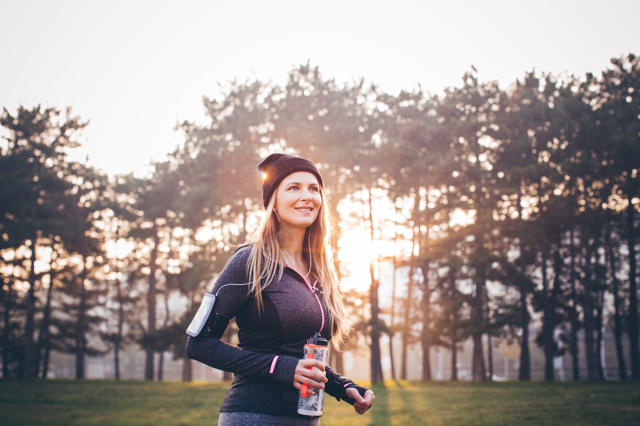 Young woman jogging outside in sunny autumn forest. She took a break to rest and drink water. The sun is shining in the background .