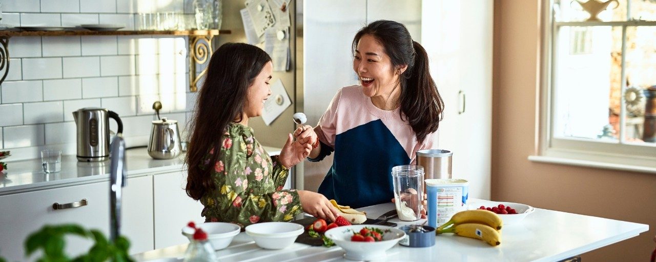 Woman holding yoghurt on spoon towards girl, laughing, threatening to smear food on daughter's face, messing about, having fun, playing, bonding