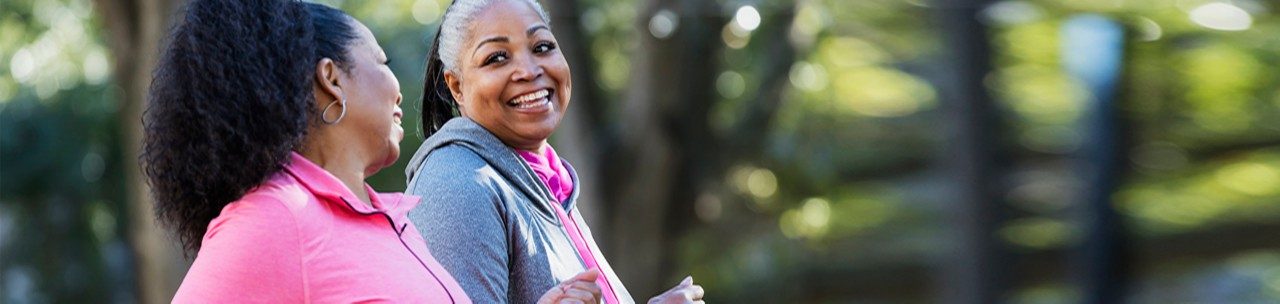 Two African-American women exercising together in the city, jogging or power walking, laughing and conversing. Buildings and trees are out of focus in the background. The one in pink is in her 60s and her friend is in her 50s.