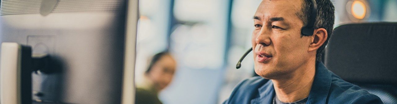 Photo of a mature Asian man with a headset using a computer while working in a call centre.