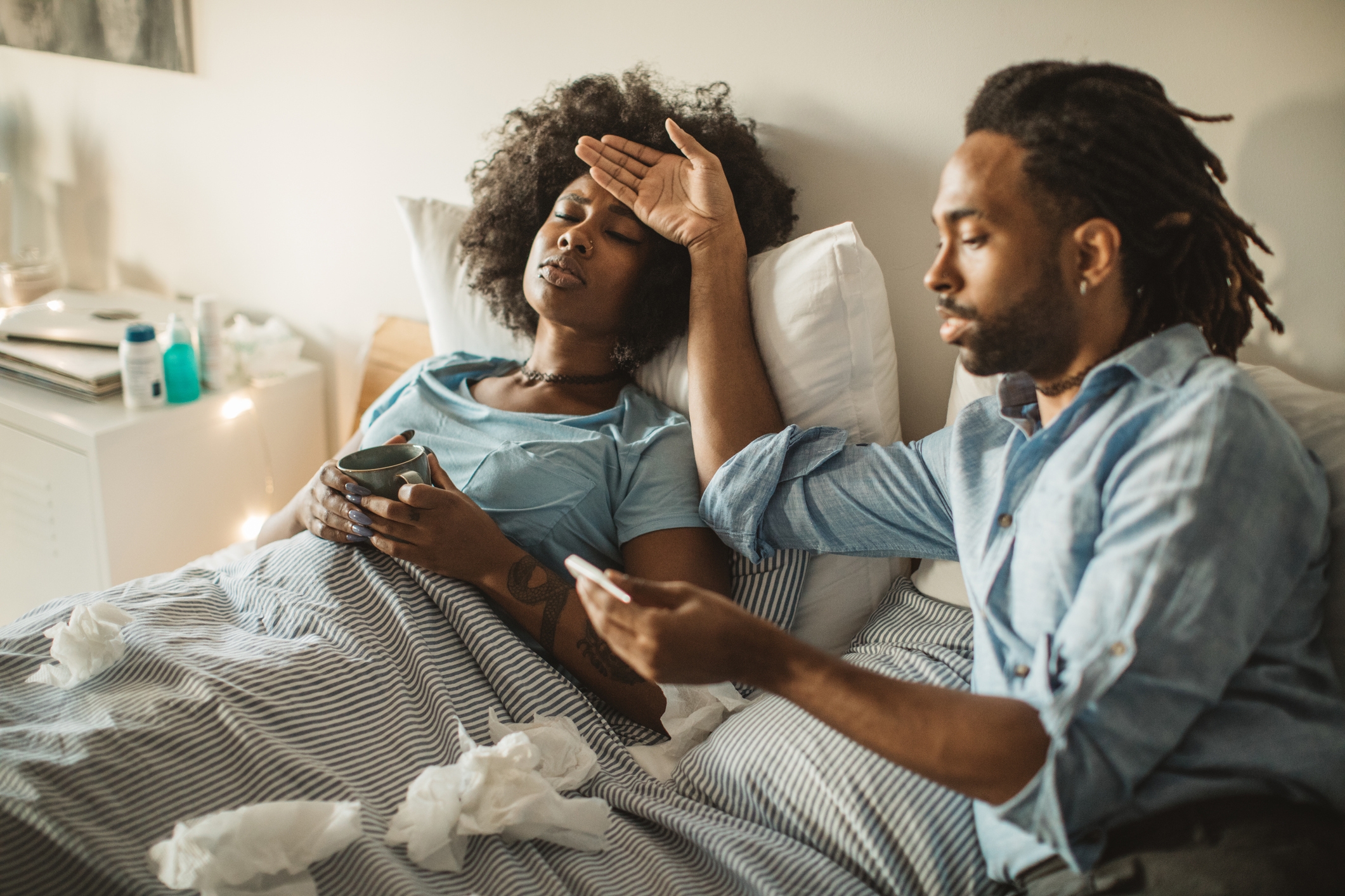 Young African-American woman feeling sick at home. Lying in bed, drinking tea and medicine. Her boyfriend or husband takes care of her. Daytime.