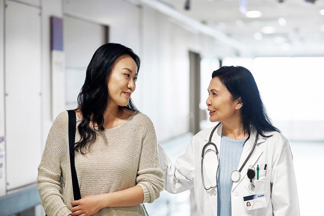 Smiling mature doctor talking to woman in hospital. Female patient visiting healthcare worker for routine checkup. They are standing in corridor.