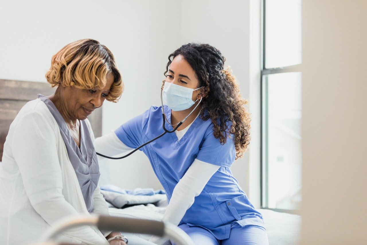 The senior adult woman sits quietly as the mid adult female nurse uses a stethoscope to check her heart and lungs during a home health visit. They both sit at the foot of the patient's bed.