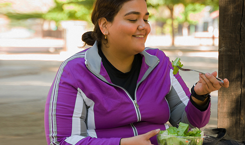 Happy overweight woman smiling while eating a healthy green salad before starting her cardio workout outdoors