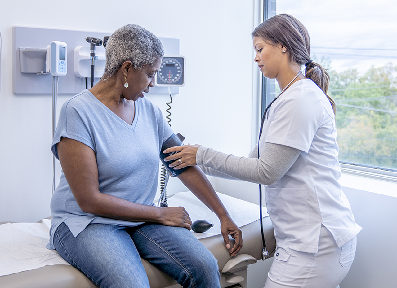 A senior patient of African decent sits up on an exam table as her female doctor takes her blood pressure. She is dressed casually and looking down at her arm as the doctor holds the blood pressure cuff.