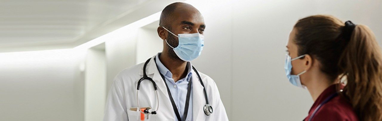 Male healthcare worker in protective mask discussing with nurse. Frontline professionals are standing in corridor during COVID-19 pandemic. They are in hospital during state of emergency.